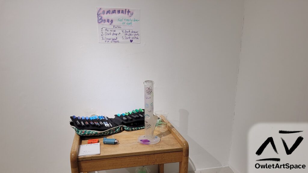 In the corner there is a wooden coffee table. Above it is a paper sign made with paint and glitter pen. It says: "Community Bong. Feel free to draw or sign!! Rules: 1. Be nice 2. Don't drop it 3. Draw small for others 4. Don't draw on clear parts 5. Don't snitch" and ends with a small heart. On the table is a frosted tall glass bong with many small drawings. Behind it rests a bag of paint pens laid out. To the left is a page titled "Pen Purge Page" with several scribbles on it. With that, a lighter and a blue albuterol asthma inhaler. There are disposable cups scattered on the table.