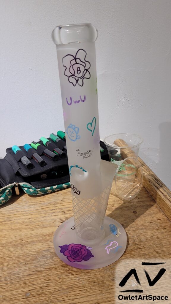 The Community Bong, resting on the coffee table, is front and center. Behind is a bag of paint pens laid out and a few scattered disposable cups. On the bong, there are several drawings. Going from top to bottom: A flower with the letter B in the center An "UwU" A green outline of a heart A small blue face "Sarah K" with hearts on each side A blue signature An anglerfish A pink signature A red rose with maroon petals on each side A pink fish outline with waves around it A yellow and blue hedgehog