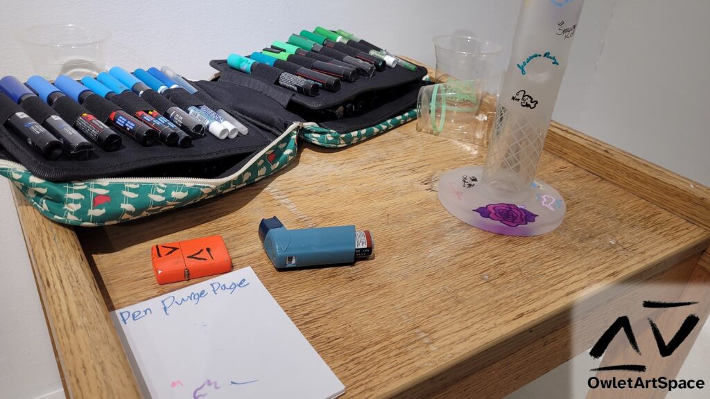 A close up of the coffee table on which is the Community Bong, featured in the left. It shows a white piece of paper titled "Pen Purge Page". Behind that is a lighter and a blue albuterol asthma inhaler. Behind that is a bag of paint pens laid out. On the table are scattered disposable cups.