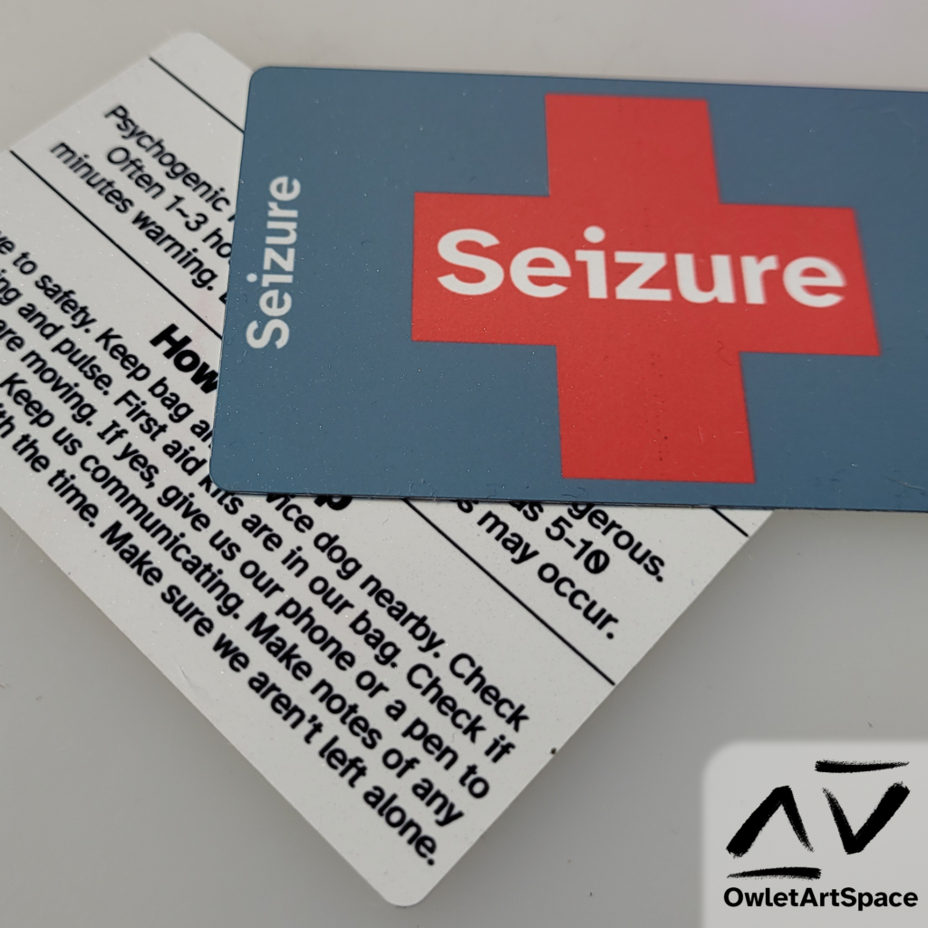 Two identical metal cards that on one side has a red cross with the word "Seizure", once in the center and two on each side. On the other side, there is the word "Seizure" on the top, then a description of the type of seizure, and then instructions on how to help.