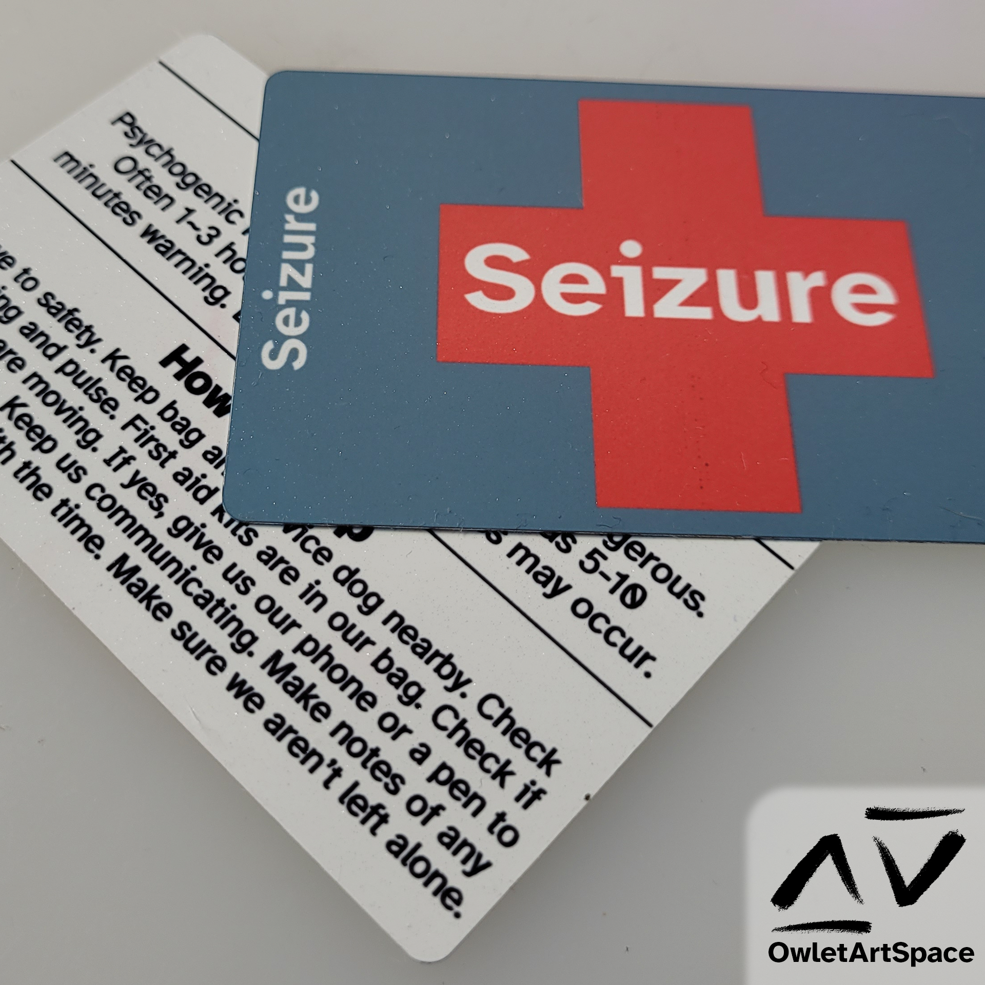 Two identical metal cards that on one side haS a red cross with the word "Seizure", once in the center and two on each side. On the other side, there is the word "Seizure" on the top, then a description of the type of seizure, and then instructions on how to help.