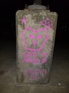 A pink owl drawing on a concrete lamp post base. Around the owl is a star, a tag, the words "Still Alive", a semicolon, and the number 741-741 (The number for Crisis Text Line)