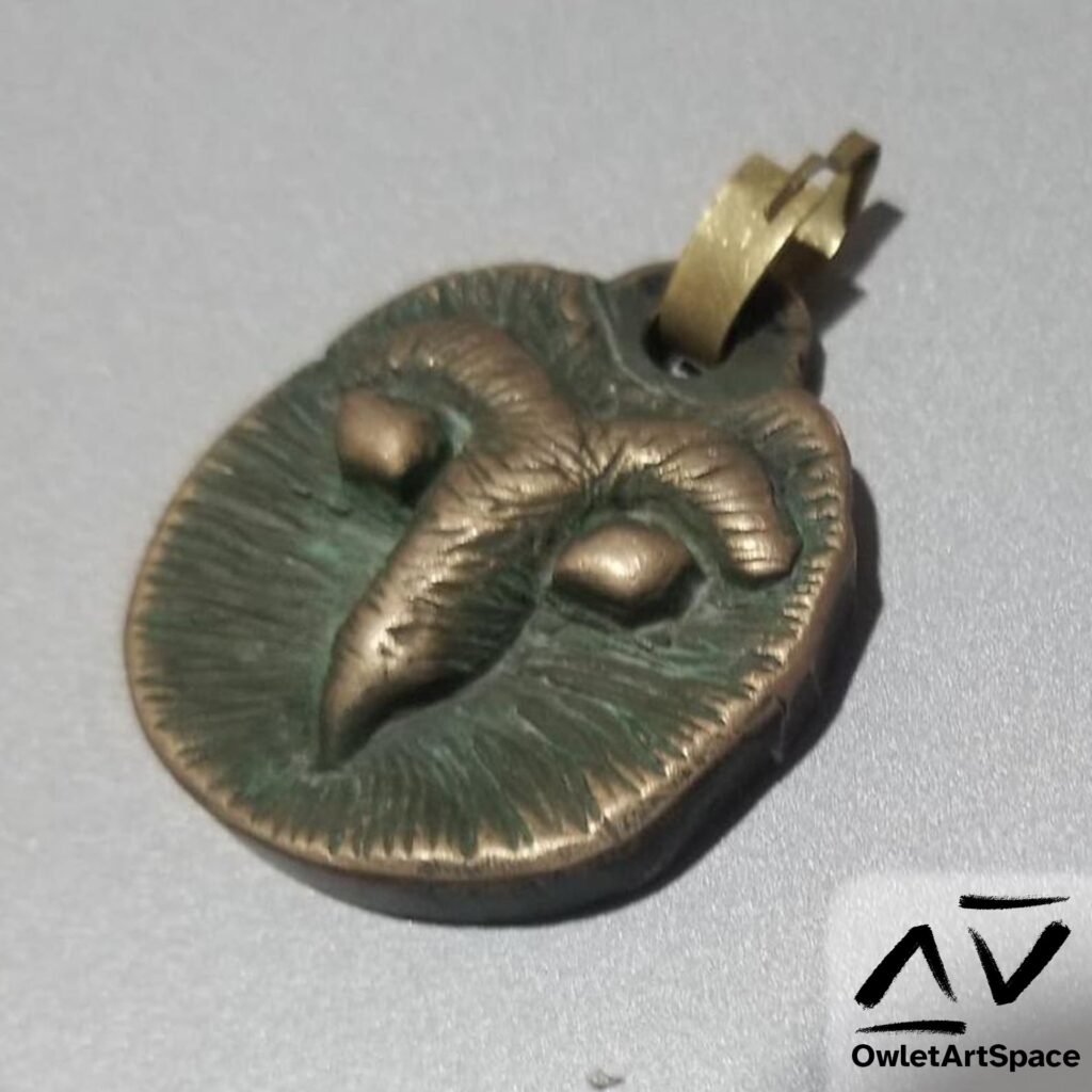 An oval pendant that has the face of a barn owl. It is made of brass. The eyes, beak, and eyebrows are raised. The rest of the face is a patina green with striations around the face. On the top is a brass clip to attach to a necklace.