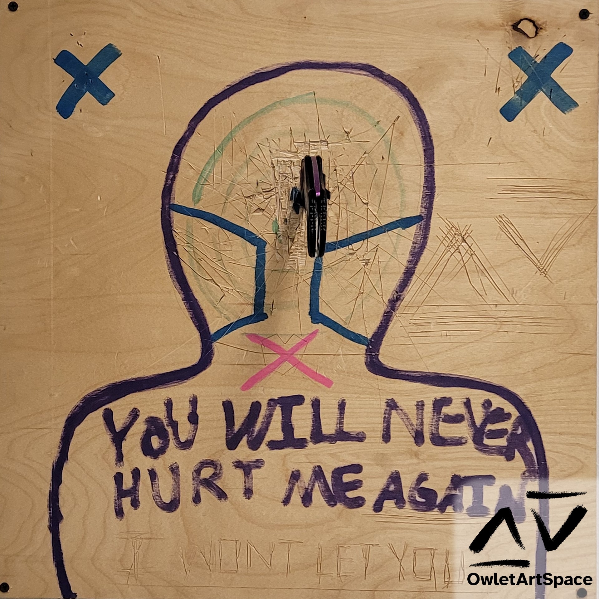 A wooden board with a silhouette with different target areas on it. The shoulder area reads "YOU WILL NEVER HURT ME AGAIN". Underneath that carved into the wood is "I WONT LET YOU" Along the main body are hundreds of knife slashes and nicks from throwing knives on two X-shaped spots above the silhouette on the left and right side. In the center of the head is a karambit sticking out of the wood.