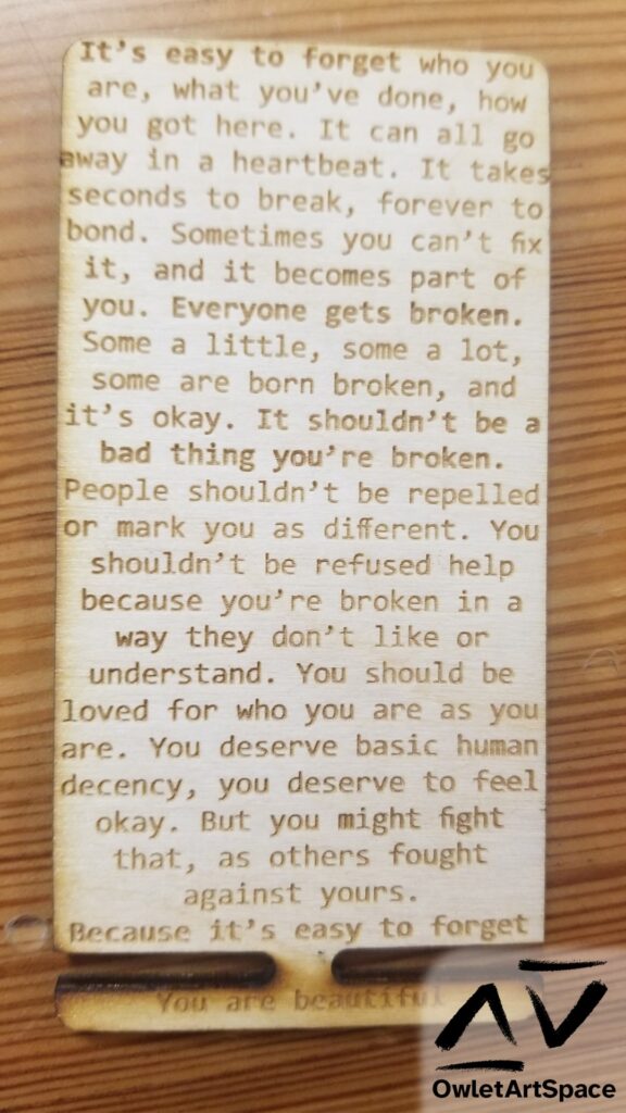 A lasercut rectangle with rounded corners. There are two sections, a large top section and a skinny section held on by a small center piece that is easy to break off. Both contain text. On the large section, it says: "It’s easy to forget who you are, what you’ve done, how you got here. It can all go away in a heartbeat. It takes seconds to break, forever to bond. Sometimes you can’t fix it, and it becomes part of you. Everyone gets broken. Some a little, some a lot, some are born broken, and it’s okay. It shouldn’t be a bad thing you’re broken. People shouldn’t be repelled or mark you as different. You shouldn’t be refused help because you’re broken in a way they don’t like or understand. You should be loved for who you are as you are. You deserve basic human decency, you deserve to feel okay. But you might fight that, as others fought against yours. Because it’s easy to forget" On the small section, it says: "You are beautiful"