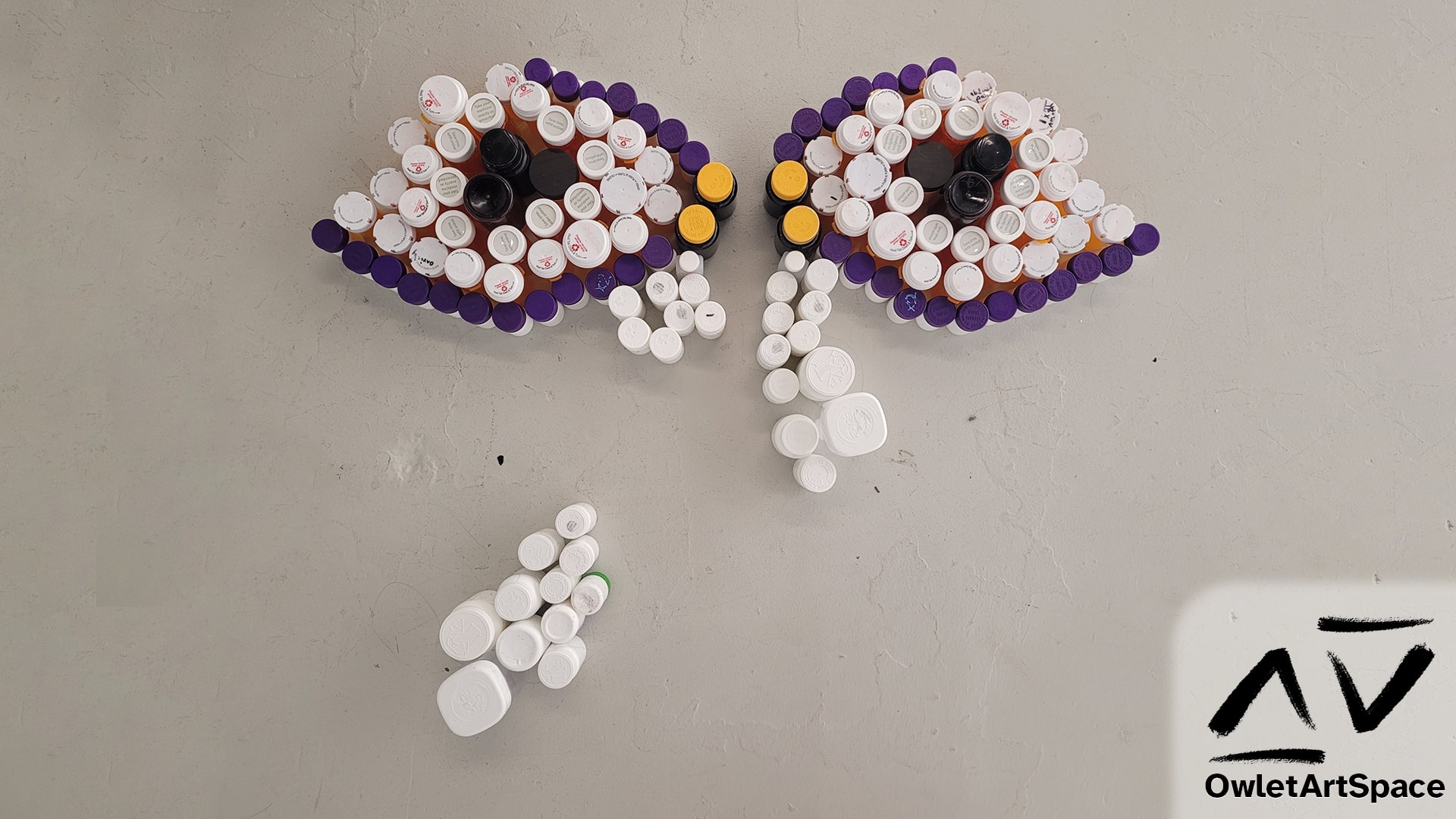 Multiple colors of pill bottles arranged to create a sleep-deprived crying set of eyes. It has black pupils, a white and orange sclera, yellow tear ducts, and white tears dripping down.