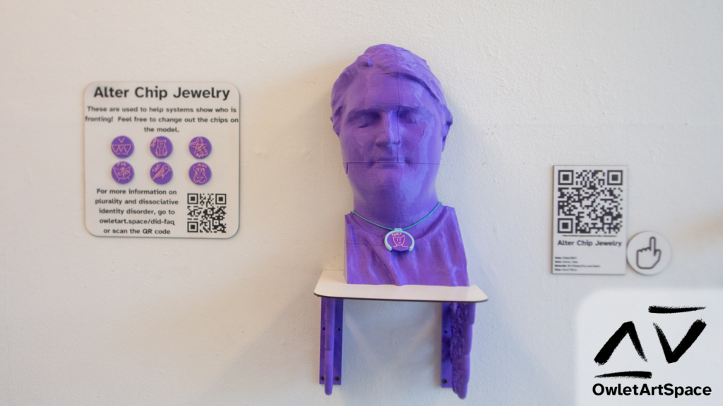 The alter chip necklace on a purple head model on a shelf. On the right is a wall label with a QR code to the page you are currently reading. On the left is a display with six alter chips held on with magnets. It reads "Alter Chip Jewelry. These are used to help systems show who is fronting! Feel free to change out the chips on the model. For more information on plurality and dissociative identity disorder, go to owletart.space/did-faq or scan the QR code."