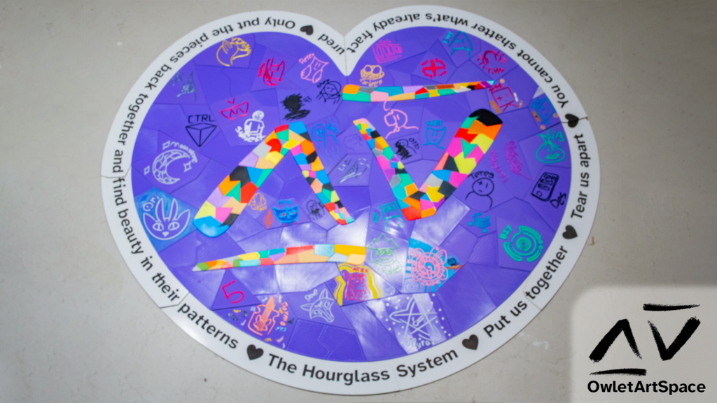 A purple heart surrounded by text with our system's symbol in the middle. The text reads "Put us together - Tear us apart - You cannot shatter what's already fractured - Only put the pieces back together and find beauty in their patterns - The Hourglass System". The center symbol is fractured and made up of different colors. The purple heart is constructed of different plates. Some are blank, the rest have the symbols of our different alters. Some of them are decorated by those alters.