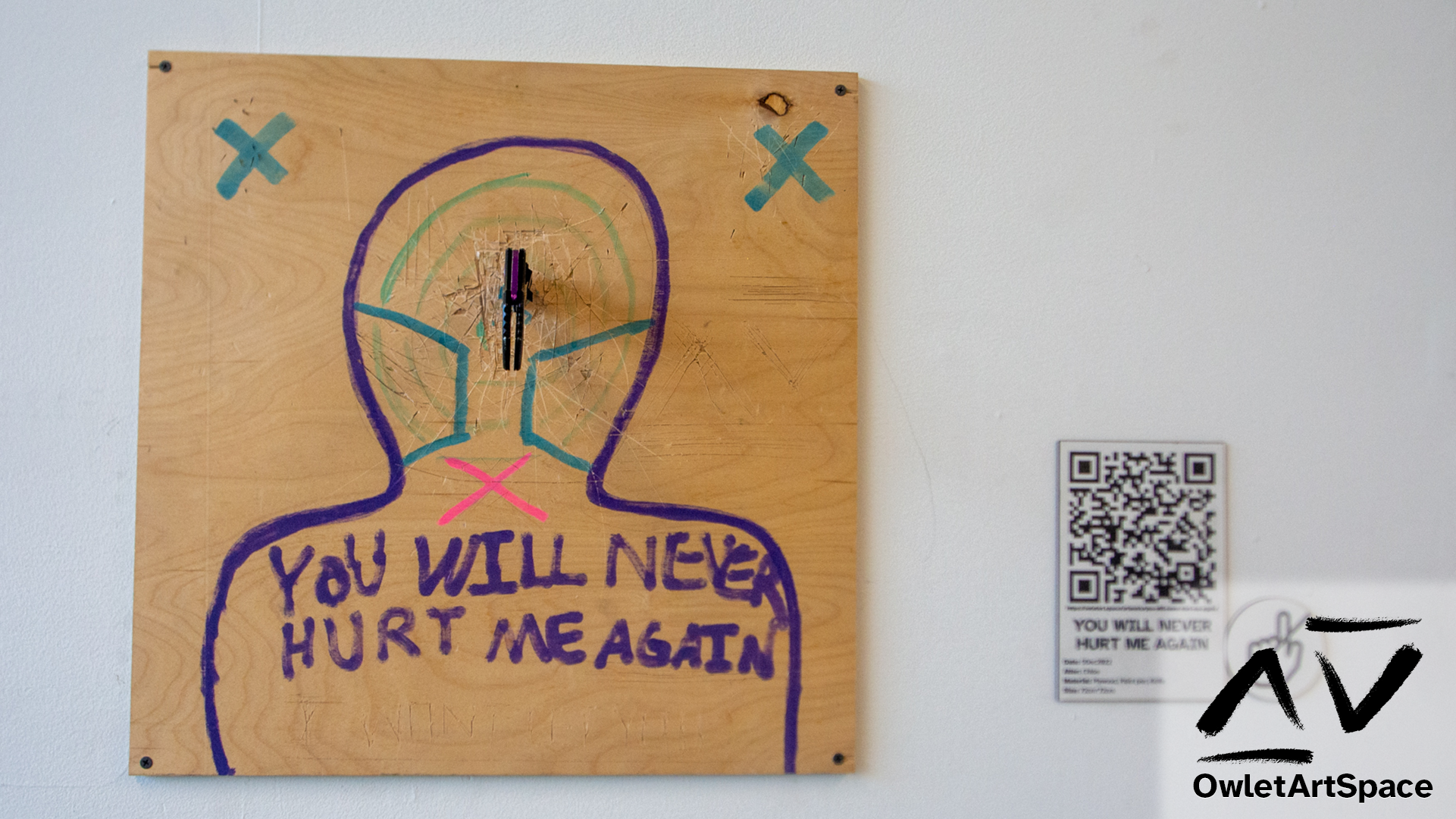 The piece YOU WILL NEVER HURT ME AGAIN. Next to it is a wall label with a QR code to the page you are currently reading.